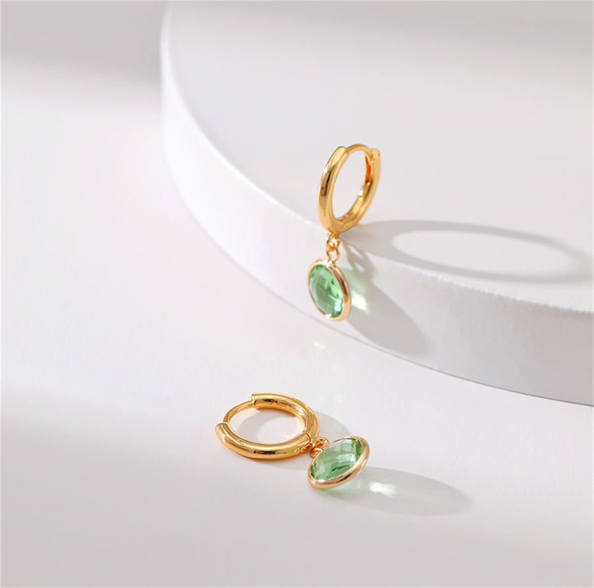 A pair of round huggie earrings in 18K gold plating, embellished with light green crystals for a chic appearance.